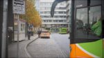 Ein Bus fährt in den zentralen Omnibusbahnhof (ZOB) ein. Im Hintergrund steht Herr Werdermann mit einem Zettel in der Hand, bereit, die Einfahrtsgebühr zu kassieren. Herbstliche Bäume und Gebäude rahmen die Szene ein.