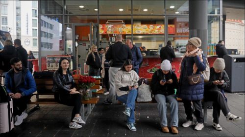 Wartende Menschen am zentralen Omnibusbahnhof (ZOB), im Hintergrund ein Döner-Imbiss. Im Vordergrund sitzt eine junge Frau mit einem großen Rosenstrauß, eine Ukrainerin, die ihre Schwester nach vier Jahren wiedersehen wird.