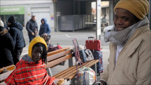 Zwei Artisten aus Kenia am zentralen Omnibusbahnhof (ZOB). Einer der Männer isst einen Döner, während der andere direkt in die Kamera blickt. Im Hintergrund sind wartende Reisende zu sehen.