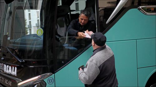 Herr Werdermann steht am geöffneten Fahrerfenster eines Busses und überreicht dem einfahrenden Busfahrer eine Quittung über die Einfahrtsgebühr von 14 Euro, während der Busfahrer bezahlt.